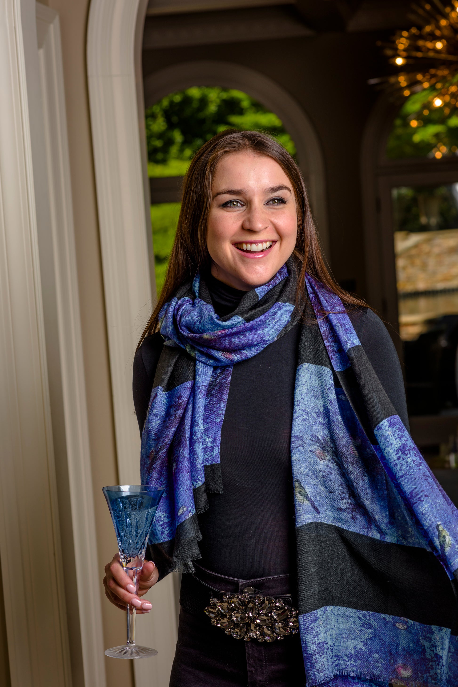 A woman standing in an elegant room wearing a multicolored cashmere silk scarf with a geometric pattern, smiling and holding a glass.